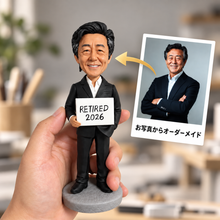 画像をギャラリービューアに読み込む, 手のひらに乗せたオーダーメイドフィギュアの実物写真。黒いスーツを着た男性が「RETIRED 2026」と書かれたプレートを持つ姿を再現。右側には参考写真と「お写真からオーダーメイド」の文字があり、退職記念や還暦祝いのプレゼントに最適な一品であることを示しています。