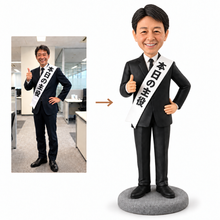 画像をギャラリービューアに読み込む, 写真をもとに製作したビジネススタイルのオーダーメイドフィギュア。黒いスーツとネクタイを着用し、「本日の主役」と書かれた白いたすきをかけています。足元にはグレー台座を配置。退職記念や還暦祝い、誕生日プレゼントに最適な一品。
